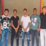 Eight men stand in a row indoors; several have yellow restraints around their wrists or hands tied with yellow bands.