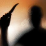 Blurred silhouette of a person with a raised knife against an orange-tinted background.