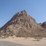 Desert landscape with a large jagged rocky hill under a clear blue sky; sparse trees and a small cluster of buildings at the base of the hill.
