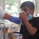 Boy sits in a chair while a clinician in full PPE holds an oxygen mask to his face, assisting his breathing.