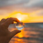 Close-up of a hand holding a glass marble at sunset, reflecting the sun and ocean in the sphere.