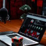 Podcast recording setup: a MacBook laptop on a desk with a colorful Rubik's cube in front, and a large microphone to the left in a red-lit studio.