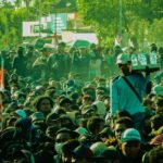 Dense crowd participating in an outdoor protest, with a man in a teal shirt and white cap carrying a water rack on his shoulder