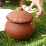 Brown clay pot with a lid sits on green grass, with hands nearby wiping or handling a cloth.
