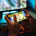 Person plays a mobile game on a smartphone, with a colorful gaming setup and RGB keyboard in the background.