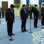 Group of men in suits and traditional caps bowing at mic stands during an official ceremony in a marble-filled room with ornate decor.
