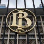 Gold circular monogram (LB) centered on a black gate with a modern building in the background.