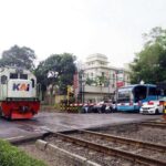 Locomotive approaching a crossing with parked buses and cars nearby at a railway intersection.