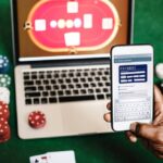 Person holds a smartphone showing an online payment form above a laptop on a green casino table with scattered poker chips