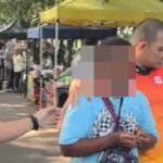 Man in a white shirt hands something to a person with a pixelated face at an outdoor market stall row under tents sunlit scene