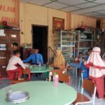 School cafeteria with round green tables, children in red-and-white uniforms eating; trophy cases along the wall to the left.