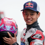 Motorsport rider in a white racing suit with red and black accents, smiling while holding a pink and blue helmet with sponsor logos, Red Bull cap in the background.