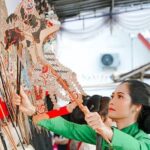 Woman in green adjusts ornate wooden shadow puppets on a display wall.