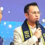 Man in a beige suit speaks into a microphone on a blue-stage backdrop, wearing a black-and-yellow scarf; another man in blue stands nearby during a talk.