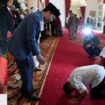Portrait of a smiling woman in a red shirt on the left; on the right, people in formal attire bowing/kneeling on a red carpet during a ceremonial event in a grand room.