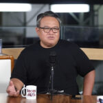Man in a black t-shirt speaks into a studio microphone at a podcast setup, with mugs on the table in the foreground and awards visible in the background.
