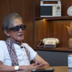 Older man with sunglasses sits at a desk in a wood-paneled office, talking during an interview.