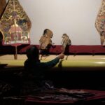 A puppeteer in a dark outfit operates two ornate traditional puppets on a dimly lit stage with decorative backdrops.