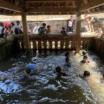 Group of children and a few adults leaning over a brick water well or canal under a wooden shelter, wading in the water below.