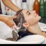 Woman reclines with head in salon sink as stylist washes her hair with shampoo bubbles present.