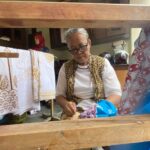 Older woman with glasses sits at a wooden loom, weaving patterned fabric as colorful textiles hang nearby, with a person in a red headscarf in the background.