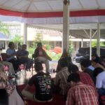 Person holding a microphone and speaking to a small audience seated in a semicircle under a red-and-white canopy outdoors, with plants in the background.