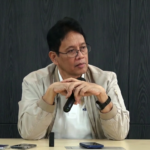 Man with glasses and a beige jacket sits at a table, holding a microphone and looking to the side as he speaks or listens.