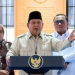 Government official in a beige uniform speaks at a podium with the Indonesian eagle crest; two aides stand behind him, flags in view.