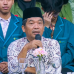 Man in batik shirt and black cap speaks into a microphone at a formal event, with students in teal blazers around him and flowers on the table.