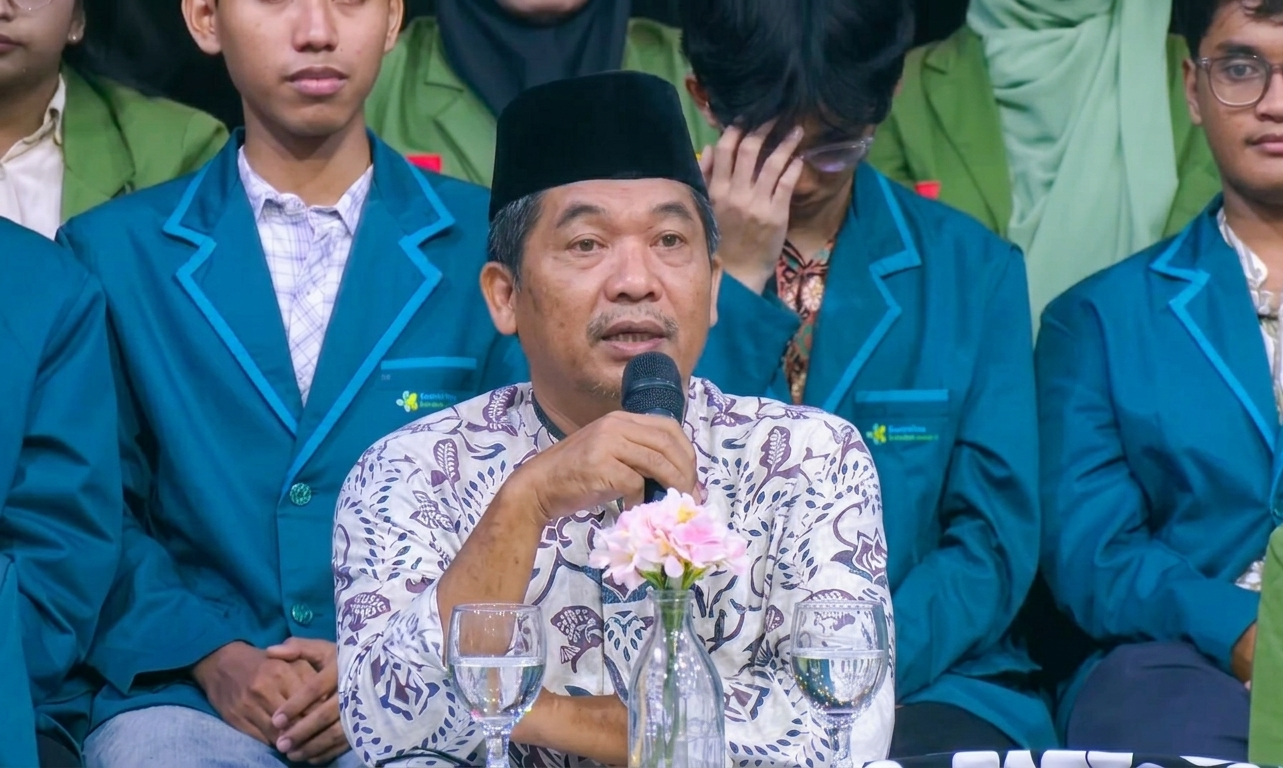 Man in batik shirt and black cap speaks into a microphone at a formal event, with students in teal blazers around him and flowers on the table.