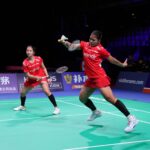 Two female badminton players in red uniforms compete on a bright green court, one reaching with her racket for a shot, shuttlecock in play.
