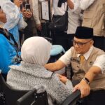 A man in a beige uniform and black cap kneels beside an elderly woman in a wheelchair, holding her hands in a crowded room. The man’s name tag reads ‘Irfan Yusof.’