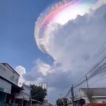 Ring-shaped iridescent cloud arc forming a rainbow circle in a blue sky above a street and buildings.