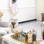 Two people sit on ornate white-and-gold couches across a small table in a bright room, casually talking; man in white cap and T-shirt on the left, two women in hijabs on the right.