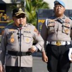Two Indonesian police officers in khaki uniforms stand outdoors; one speaks into a microphone while the other looks on.