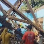 People stand amid a tangle of large wooden beams and debris from a collapsed construction structure, with a blue tarpaulin in the background and onlookers nearby.