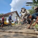 Group of workers repaving a small road in a rural neighborhood, using a concrete mixer and shovels while a few men help spread gravel and level the surface.