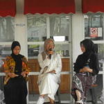 Three women wearing hijabs sit on a stage; the middle woman speaks into a microphone while the others listen or hold notes.