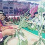Group of Indonesian people in traditional attire weaving palm leaves at a long table during a cultural ceremony under a tent with a banner behind them.
