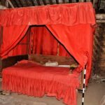 Red canopy bed with draped red curtains in a rustic room, a low wooden frame and ruffled bed skirt visible.