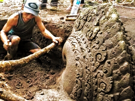 Person kneeling in mud, digging around a large ornate carved stone wheel.
