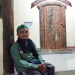 Two people sit on a white floor beside massive wooden pillars inside a hall, with a sign reading Makam Syed Djangkung above the door.