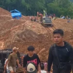 Construction site on a muddy slope with an orange excavator and workers; crowd visible in the background on the hill carven by earthworks.