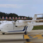 White twin-engine turboprop airplane parked on a taxiway with blue-tail livery and 'HH-100' on the fuselage, banners in the background.