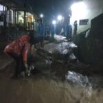 Nighttime street flooded with muddy water; people in rain gear wade through debris and mud under streetlights.