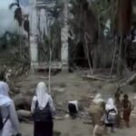 Group of people in headscarves walking through a ruined village with collapsed structures and debris in the background.
