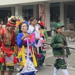 Group of costumed performers in bright outfits and face paint marching down a city street.