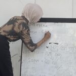 Woman in a light hijab writes on a whiteboard filled with handwritten notes in a classroom setting.