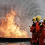 Firefighters in orange suits and yellow helmets aim a hose at large flames during a blaze.