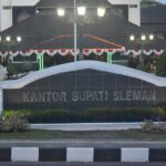 Signage at a gov’t building reading 'KANTOR BUPATI SLEMAN' behind a curved white wall with hedges and lights at dusk/uniformed garden area